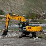 Escavadeira amarela trabalha em terreno rochoso às margens de um rio em área rural.