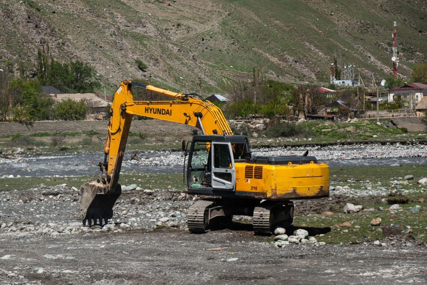 Escavadeira amarela trabalha em terreno rochoso às margens de um rio em área rural.