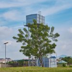 Torre de água em uma área rural, cercada por vegetação e árvores sob céu azul.