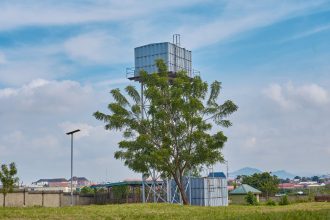 Torre de água em uma área rural, cercada por vegetação e árvores sob céu azul.