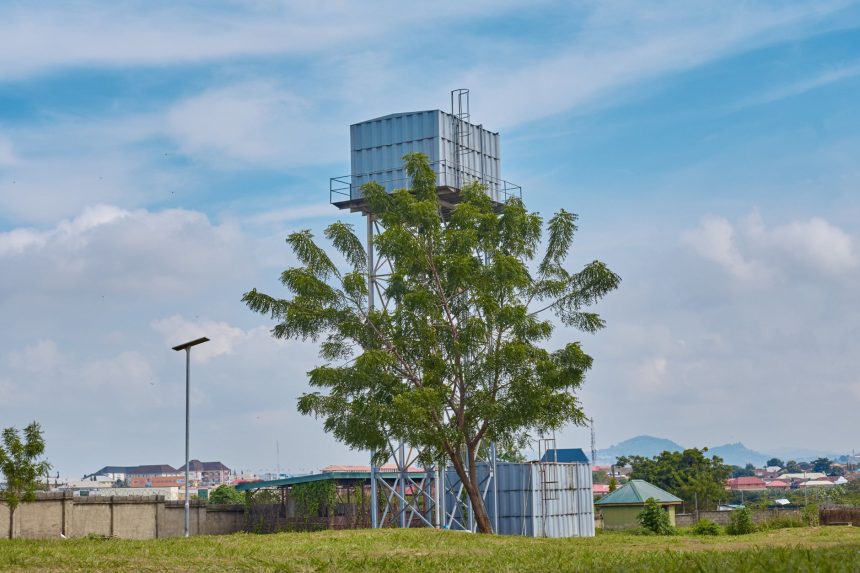 Torre de água em uma área rural, cercada por vegetação e árvores sob céu azul.