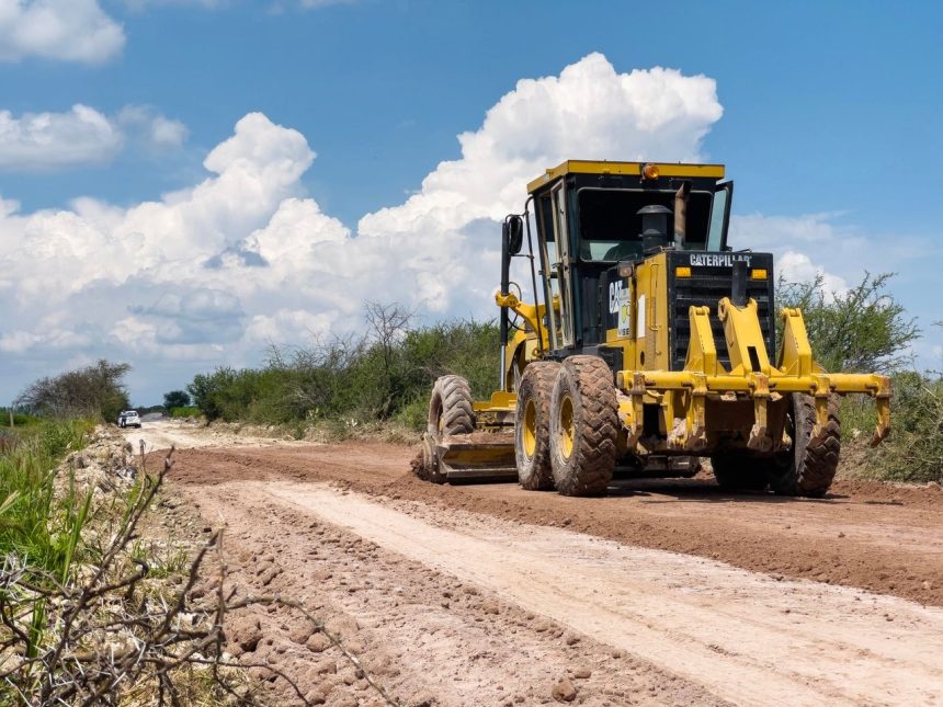 Motoniveladora amarela trabalhando no nivelamento de uma estrada de terra em área rural, sob céu ensolarado.