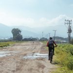 Ciclista percorre uma estrada de terra em área rural, com montanhas ao fundo e céu nublado.