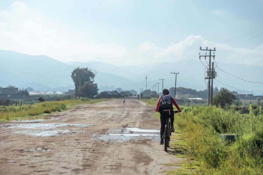 Ciclista percorre uma estrada de terra em área rural, com montanhas ao fundo e céu nublado.