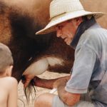 Homem idoso e criança ordenham uma vaca ao ar livre em uma propriedade rural.